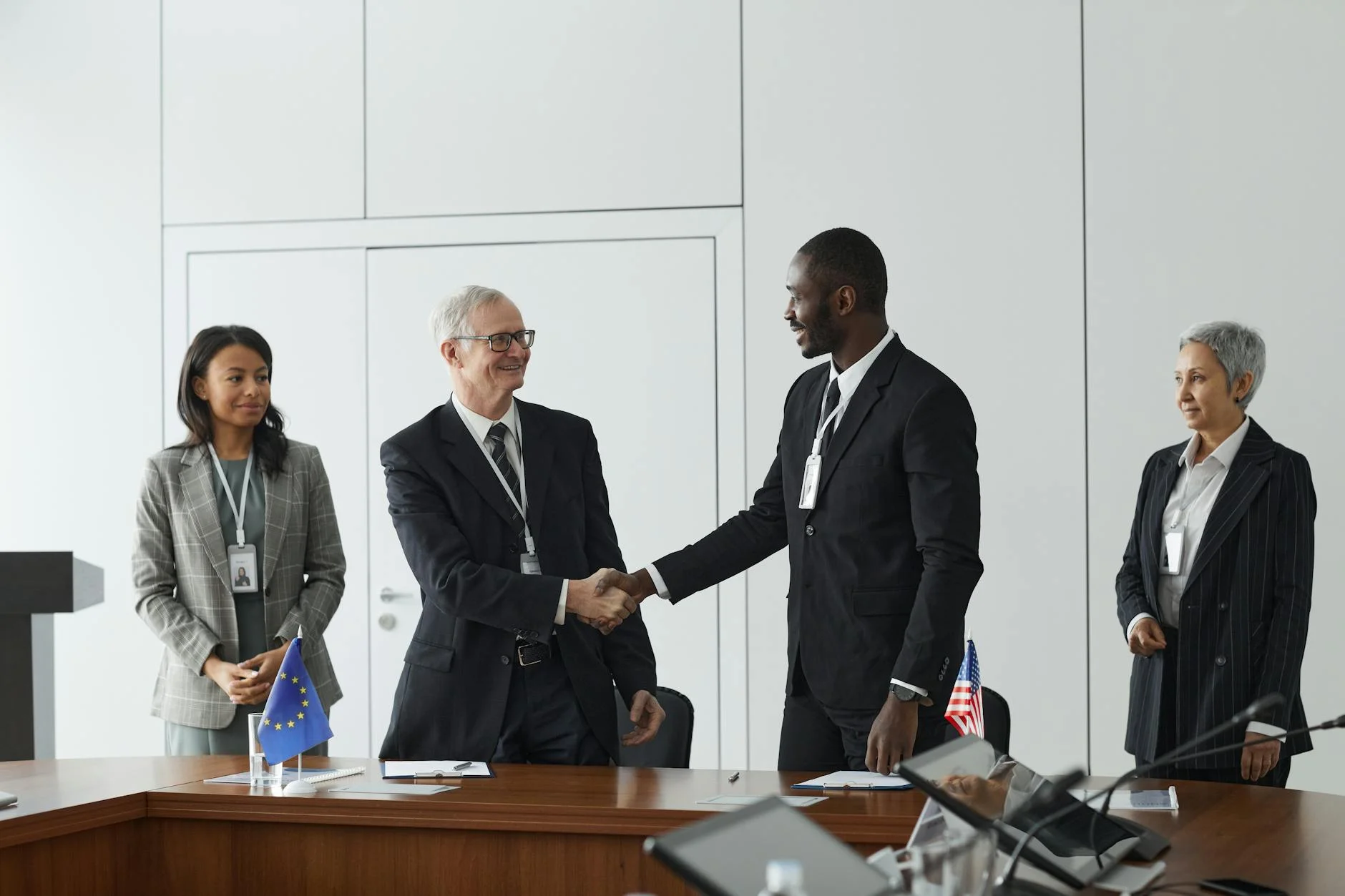 Business professionals shaking hands in conference room with flags signifying international agreement.