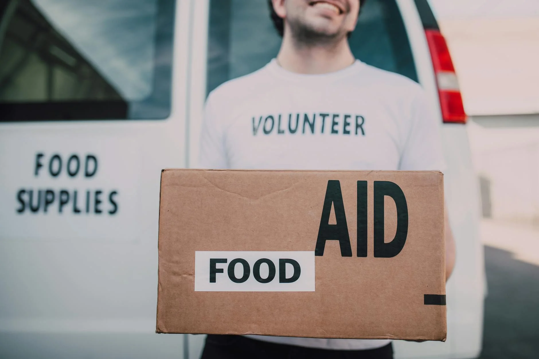 Smiling volunteer holding a box labeled 'Food Aid' outside a van with 'Food Supplies' sign.