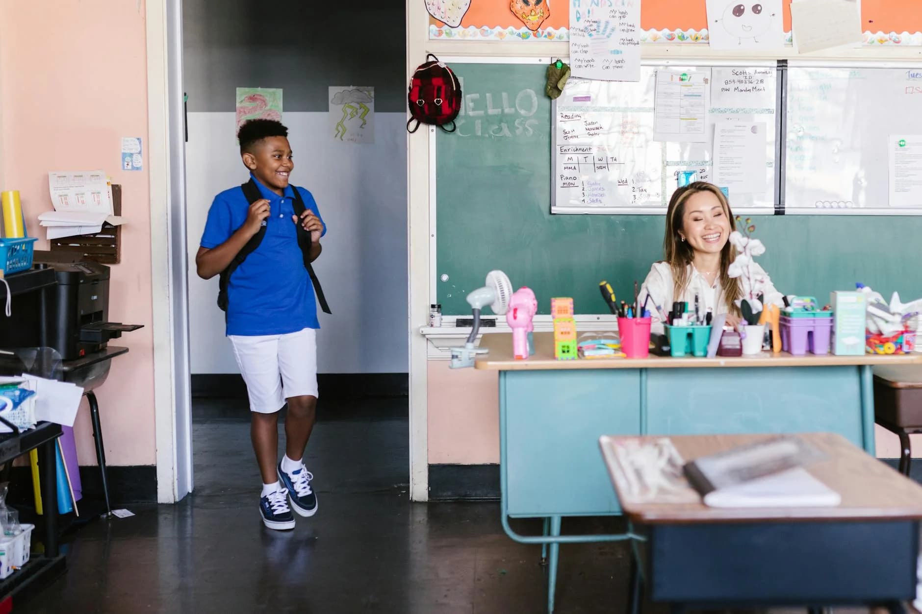 A cheerful student enters the classroom where a teacher is seated at a desk, ready for class.