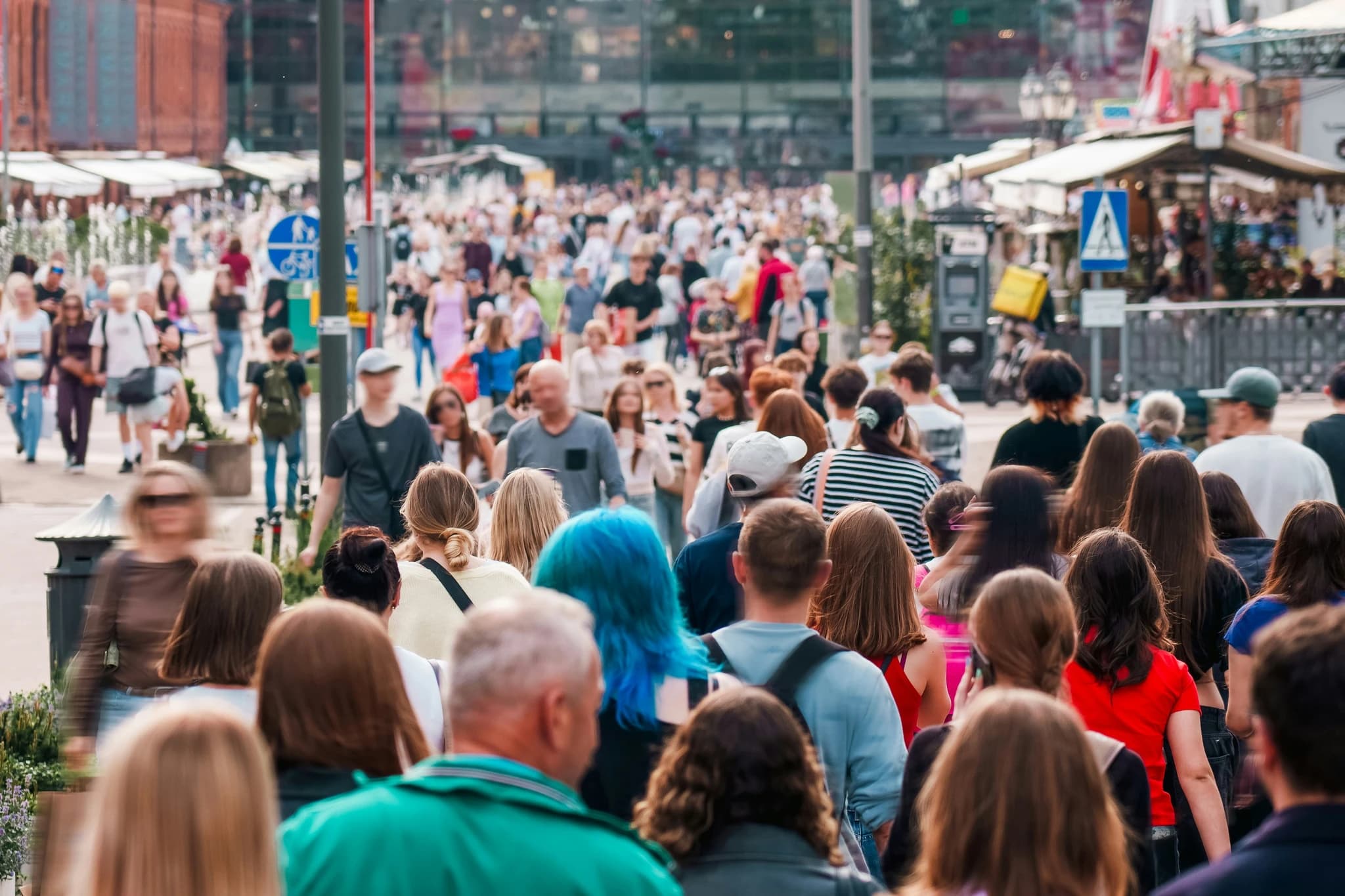 A large, diverse crowd of people walks through a sunny, open-air pedestrian plaza lined with stalls and urban infrastructure. The scene captures a lively, casual atmosphere, with the motion blur of nearby pedestrians emphasizing the active flow of the busy outdoor space.