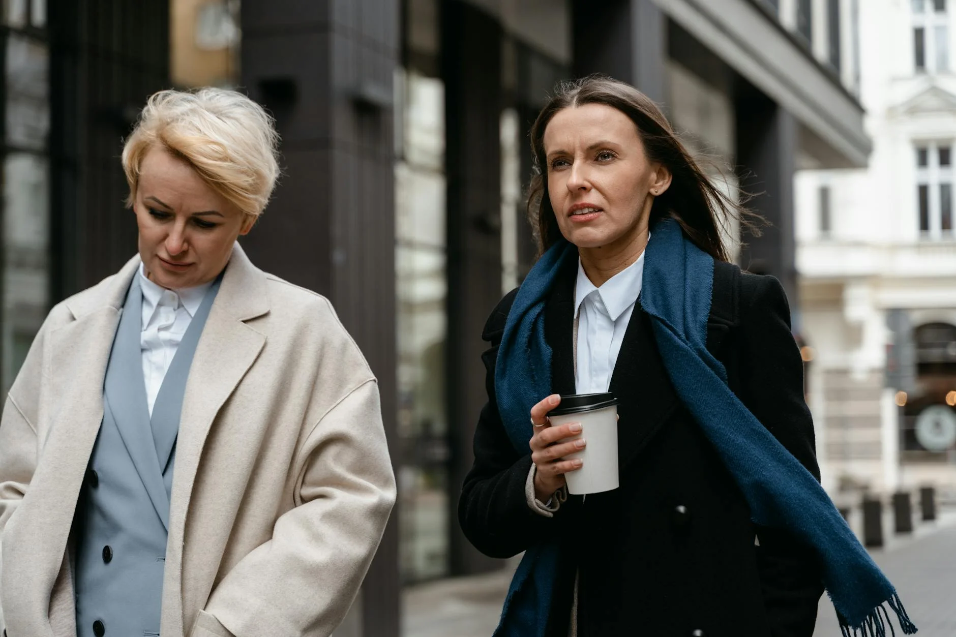 Two businesswomen in coats conversing and walking with a coffee cup on a city street.
