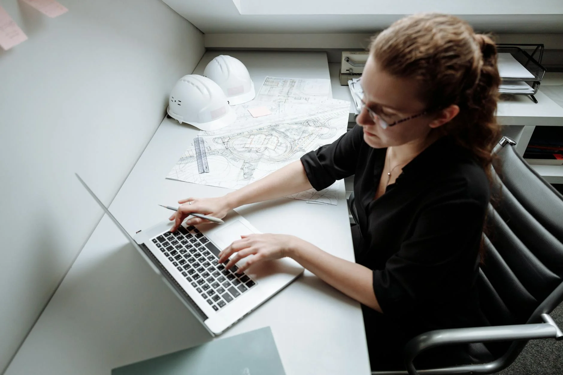 Woman architect working on a laptop at her desk with blueprints and safety helmets.