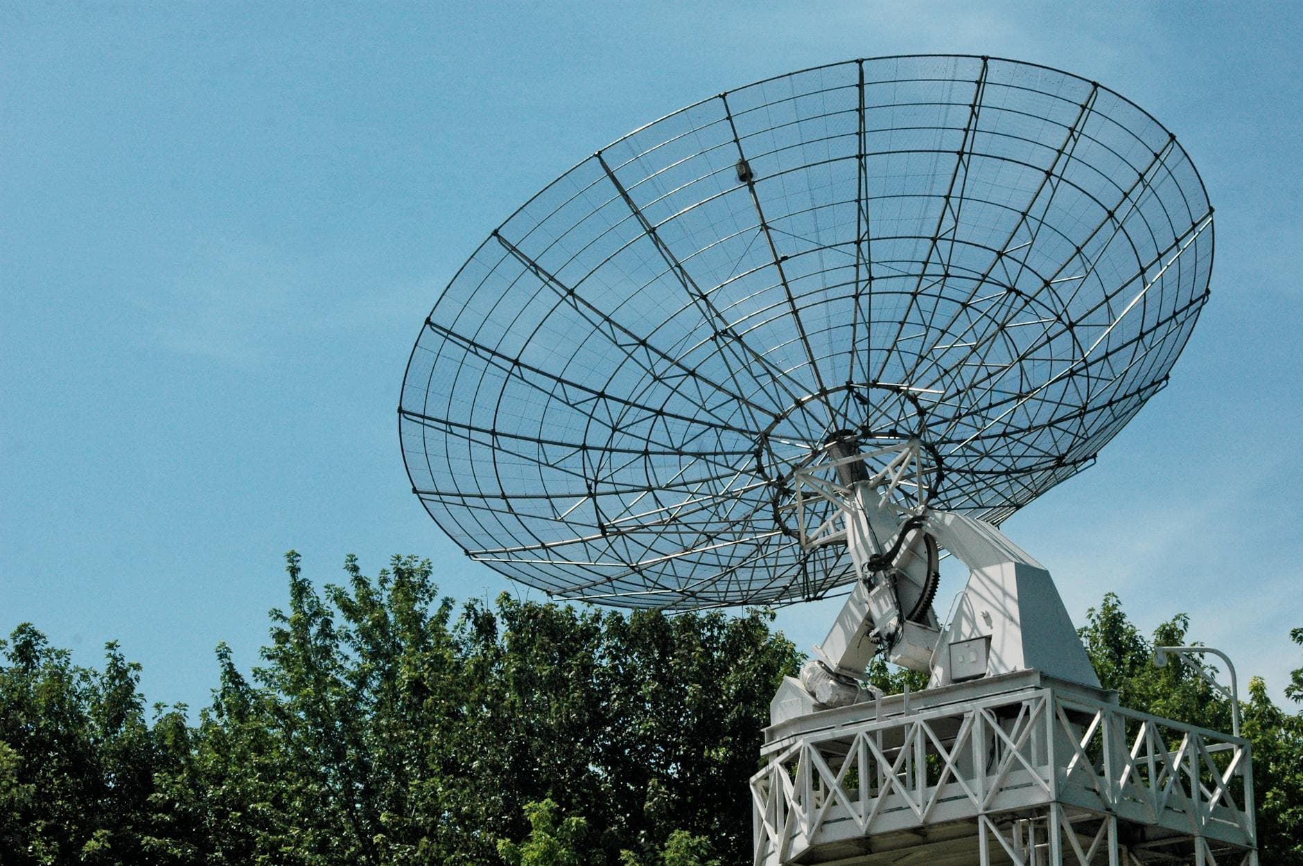 A large satellite dish stands tall against a clear blue sky, surrounded by greenery.