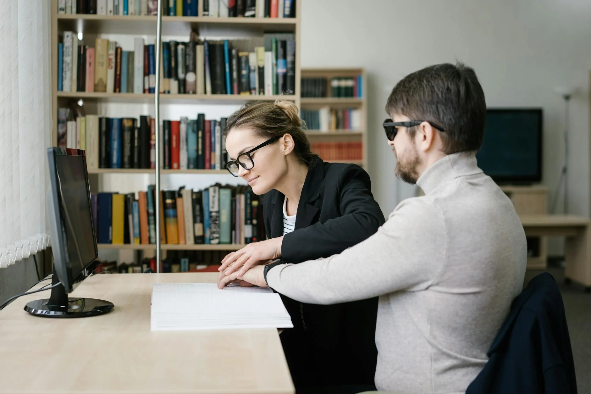 A caregiver helps a visually impaired man read Braille in a library setting.