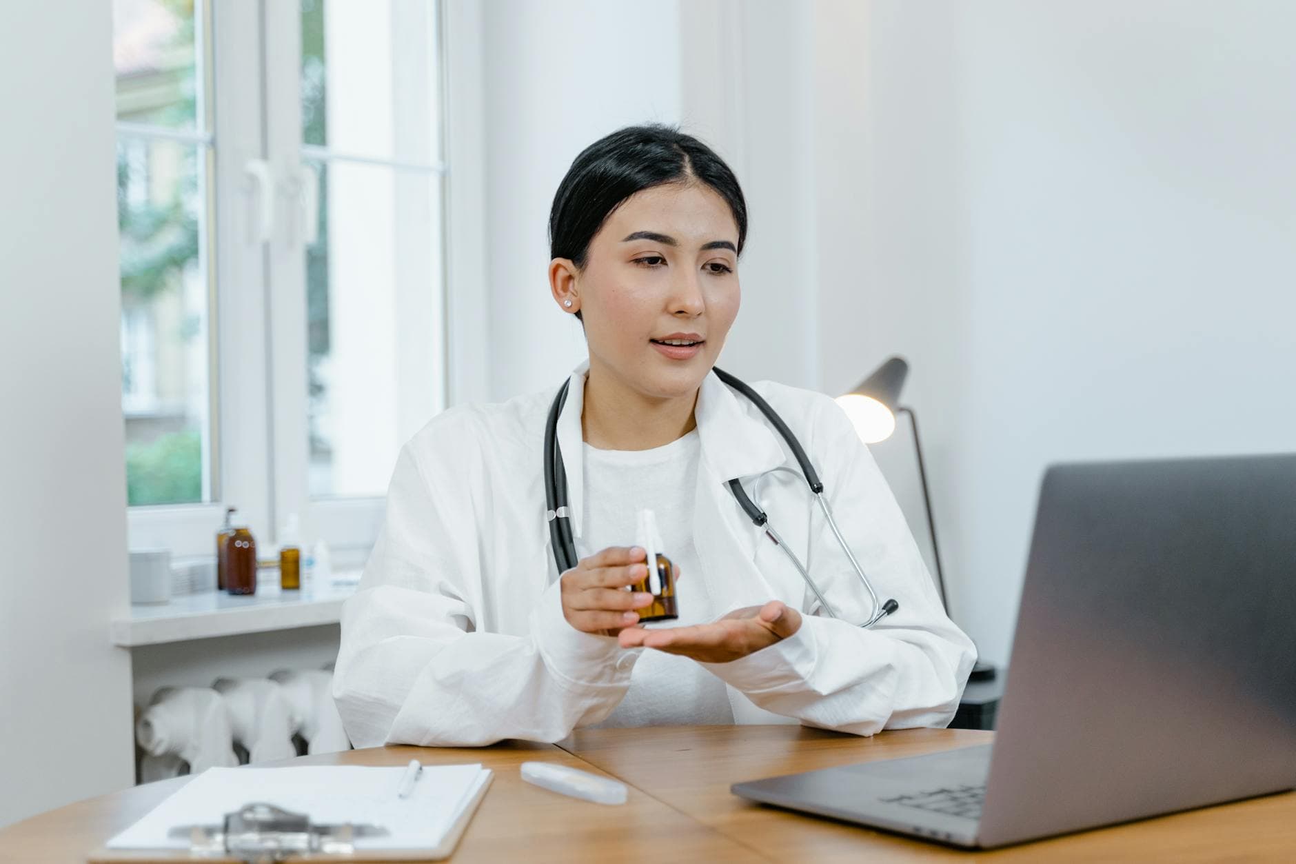 A young female doctor in a white coat on a video call, discussing medication with a patient.