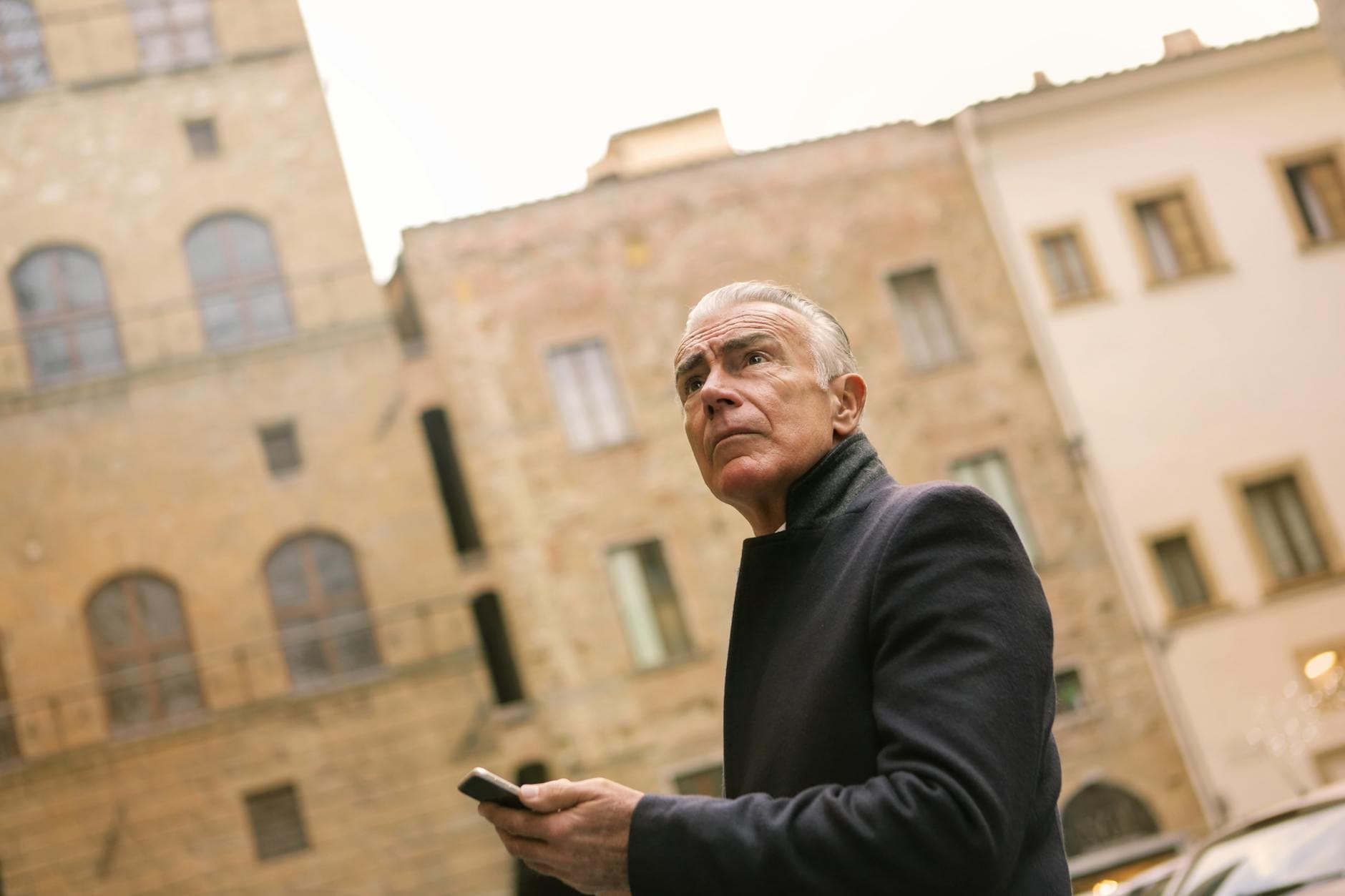 Senior man outdoors holding a phone with historical building background.