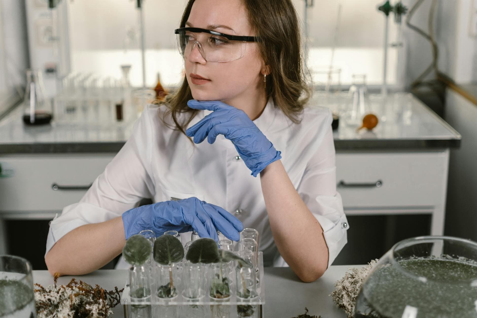 A female scientist wearing protective gear examines plant samples in a laboratory setting.
